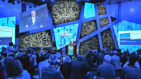 Begehrter Redner auf der SCCON 2025: Bundesdigitalminister Dr. Karsten Wildberger Blick über die Zuschauerreihen auf die Hauptbühne der SCCON 2025, wo Bundesdigitalminister Dr. Karsten Wildberger am Redepult steht